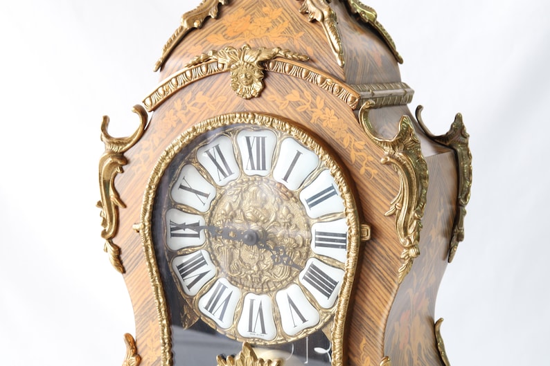 May include: A gold and brown ornate clock with Roman numeral markings on the face. The clock has a decorative design with intricate details and a glass cover over the face.