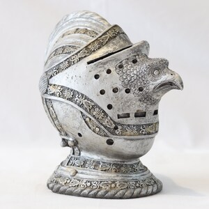 May include: A silver knight's helmet with an eagle head design. The helmet is decorated with intricate details and has a textured surface.