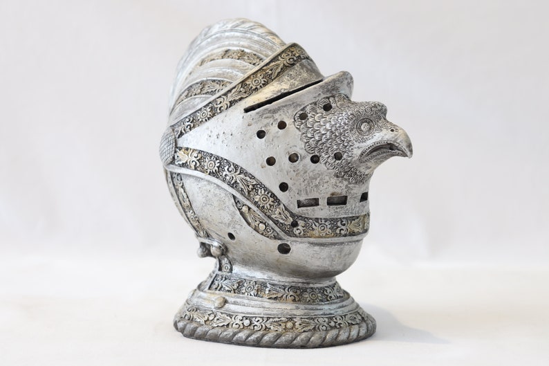 May include: A silver knight's helmet with an eagle head design. The helmet is decorated with intricate details and has a textured surface.