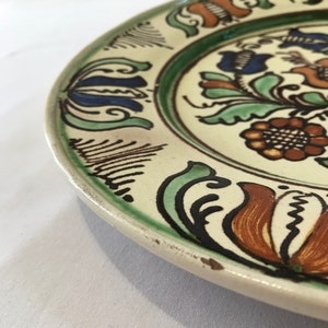 May include: A white ceramic plate with a floral design. The plate has a green, blue, orange, and brown floral pattern. The plate is slightly chipped on the edge.