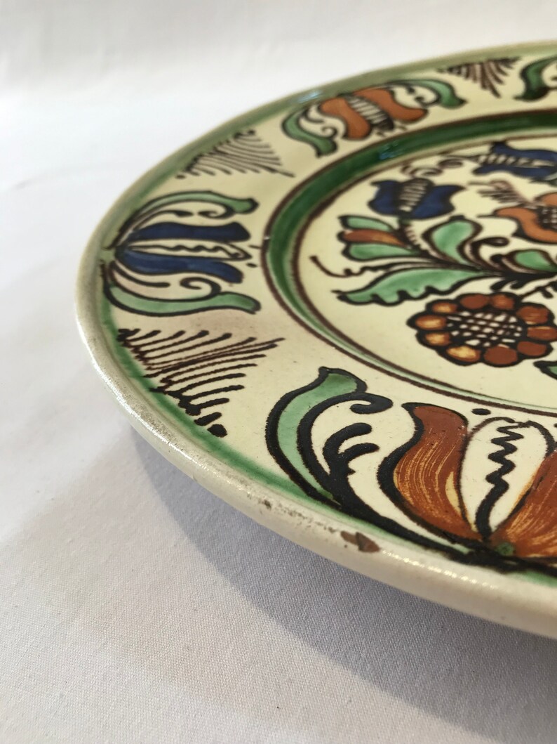 May include: A white ceramic plate with a floral design. The plate has a green, blue, orange, and brown floral pattern. The plate is slightly chipped on the edge.