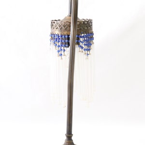K&ouml;nnte beinhalten: Eine Vintage-Tischlampe mit einem Messingfu&szlig; und einem klaren Glasschirm. Der Schirm ist mit blauen Perlen und klaren Glastropfen verziert.