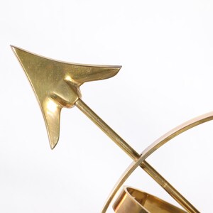 May include: A gold-toned metal sculpture of a compass with a pointed arrow. The arrow is pointing to the right.
