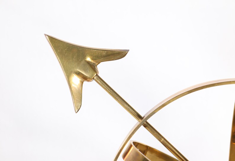 May include: A gold-toned metal sculpture of a compass with a pointed arrow. The arrow is pointing to the right.