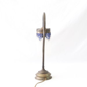 K&ouml;nnte beinhalten: Eine Tischlampe aus Messing mit einem gebogenen Arm und einem Perlenschatten. Der Schatten ist mit blauen und klaren Glasperlen verziert.
