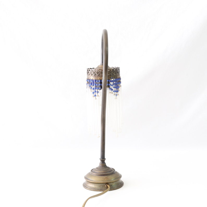 K&ouml;nnte beinhalten: Eine Tischlampe aus Messing mit einem gebogenen Arm und einem Perlenschatten. Der Schatten ist mit blauen und klaren Glasperlen verziert.