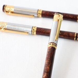 May include: Two silver and gold pens with brown wood-grain barrels. The pens have gold accents and the words "+ DUKE" are engraved on the barrel of one pen.