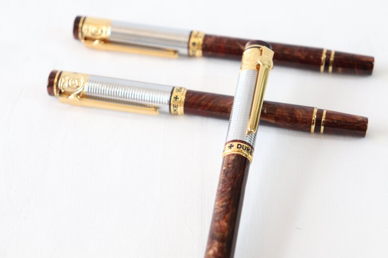May include: Two silver and gold pens with brown wood-grain barrels. The pens have gold accents and the words "+ DUKE" are engraved on the barrel of one pen.