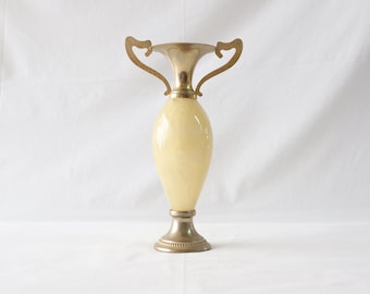 May include: A cream-colored ceramic vase with gold-tone metal handles and base. The vase has a rounded body and a flared lip.