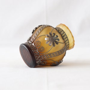 May include: A small, amber-colored glass vase with a silver metal design. The vase has a floral pattern and a geometric border.