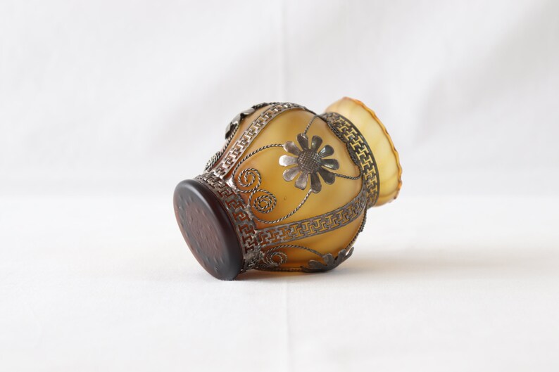 May include: A small, amber-colored glass vase with a silver metal design. The vase has a floral pattern and a geometric border.