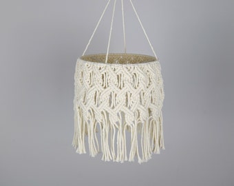 Puede incluir: Una pantalla de lámpara colgante de macramé blanca con un anillo de madera en la parte superior y una franja alrededor de la parte inferior.
