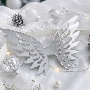 Op de afbeelding: Een zilverkleurig, reflecterend engelenvleugelornament met gedetailleerde uitgesneden veren. De vleugels zijn omringd door witte dennenappels, doorzichtige glitterballen, witte parels en een dunne lichtsnoer met warm wit licht.