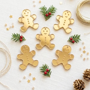 May include: Five gold-toned mirror acrylic gingerbread man ornaments, each featuring a smiling face, three circular buttons, and wavy lines on the arms and legs. Each ornament includes a small hole at the top for hanging, arranged on a white surface.