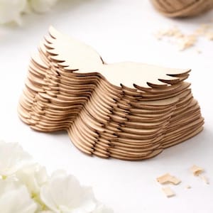 Puede incluir: Una pila de recortes de alas de ángel de madera sin terminar. Las alas son de color marrón claro con un diseño detallado de plumas. Las alas están apiladas sobre una superficie blanca con flores blancas y una bola de hilo en el fondo.