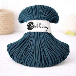 May include: A teal-colored ball of Bobbiny macrame cord. The cord is tightly wound and wrapped with a white label that has the word "Bobbiny" printed on it in a cursive font. The cord is a deep teal color, and the texture appears soft.
