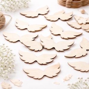 Peut inclure: Plusieurs ailes d'ange en bois non finies sont disposées sur une surface blanche. Les ailes sont de couleur brun clair, avec le grain naturel du bois. Des fleurs blanches et une pelote de ficelle suggèrent un thème artisanal ou décoratif.