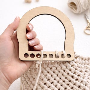 May include: A wooden bag handle with a rounded top and a series of holes for attaching to a crocheted bag. The bag is made of cream-colored yarn. The handle is held in a hand.