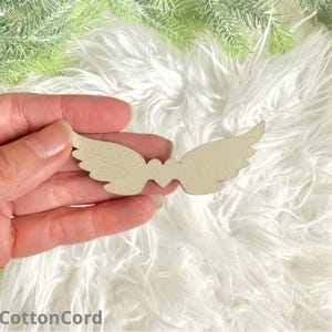 Op de afbeelding: Een paar houten engelenvleugels met een hartvormige uitsparing in het midden. De vleugels zijn beige van kleur en worden in een hand gehouden.