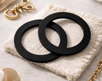 Puede incluir: Dos posavasos circulares negros se muestran sobre un cuadrado blanco roto texturizado. Una cadena dorada, un llavero y objetos decorativos están dispuestos alrededor de los posavasos sobre una superficie clara. Los posavasos tienen un diseño minimalista.