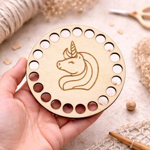 May include: A round, light-colored wooden craft base with a laser-etched unicorn design. The base has a series of evenly spaced holes around the perimeter. The unicorn has a flowing mane and a single horn. The craft base is held in a hand.