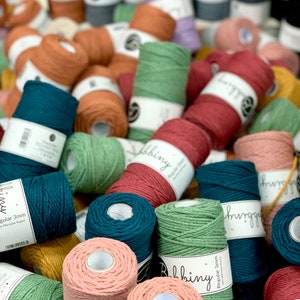 Può includere: Una varietà di rotoli di corda di cotone colorati in diverse dimensioni. I rotoli sono disposti in una pila e alcuni hanno etichette che recitano "Bobbiny" e "Remarkable 3mm".