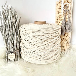 Peut inclure: Une grande bobine de corde en coton naturel, avec une étiquette en papier brun, est posée sur une surface blanche et moelleuse.  Il y a un bocal en verre rempli de perles en bois et de glands en arrière-plan.