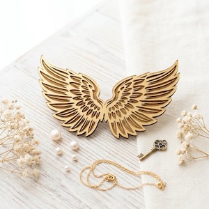 May include: A laser-cut wooden ornament shaped like a pair of symmetrical angel wings with detailed feather engravings. The wings are accompanied by a small metallic skeleton key, a delicate gold-toned chain, and several small white pearl beads.