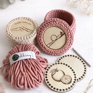 May include: A collection of handmade crochet items, including two small baskets with wooden bases, a ball of pink yarn labeled "Bobbiny", and wooden coasters with etched designs. One coaster features an orange illustration. The baskets have "Ojai Valley" etched on the wooden base.