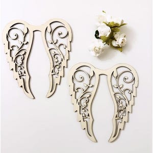 Op de afbeelding: Twee houten engelen vleugels met ingewikkelde bloemmotieven. De vleugels zijn lichtbeige van kleur en hebben een delicaat, luchtig ontwerp. Een klein boeket witte bloemen met groene bladeren is op de achtergrond.