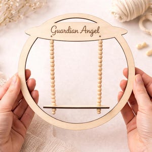 May include: A wooden Guardian Angel sign with a swing design. The sign is made of light-colored wood and features the words "Guardian Angel" in a script font. The swing has beaded strands and a small wooden seat.