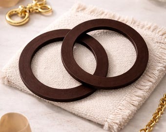 Puede incluir: Dos anillos de madera marrón oscuro descansan sobre un posavasos de lino beige. Una cadena, un llavero y una cinta dorados están dispuestos alrededor de los anillos. Los anillos tienen forma circular con un agujero en el centro.