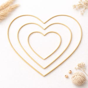 May include: Three gold-colored heart-shaped wire frames nested within each other, creating a layered effect. The frames are arranged on a white surface, with dried floral accents and a ball of twine nearby. A simple, elegant decorative item.