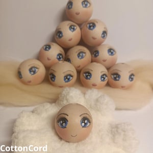 May include: A group of wooden doll heads with painted faces, including blue eyes, rosy cheeks, and eyelashes. The heads are arranged in a pyramid shape, with one head resting on a white, textured surface. The text "CottonCord" is visible at the bottom.