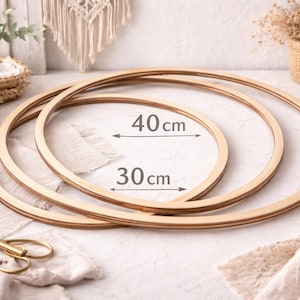Puede incluir: Se muestran tres aros de bordado de madera, de 30 cm y 40 cm de diámetro. Los aros son de color marrón claro y están apilados. La imagen incluye unas tijeras doradas, un jarrón con vegetación y otros materiales de artesanía.