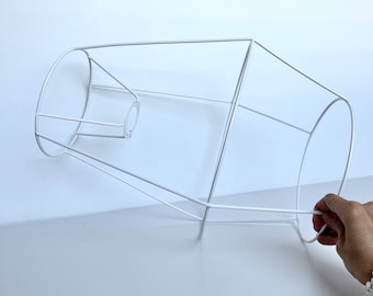 May include: White metal lampshade frame. The frame is a simple geometric shape with a circular opening at the top and a larger circular opening at the bottom.