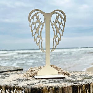 Op de afbeelding: Een houten engelenvleugel displaystandaard met een rechthoekige basis. De vleugels zijn ingewikkeld uitgesneden en de standaard staat op een verweerd houten oppervlak met zand. De achtergrond toont de oceaan en de lucht.