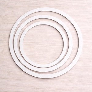 May include: Three white, circular frames of varying sizes are arranged concentrically on a light wood-grain surface. The frames appear to be made of a solid material, with a clean, minimalist design.