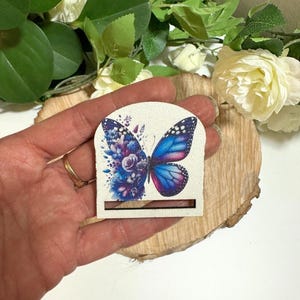May include: A decorative butterfly-shaped tag with a blue, purple, and white design. The tag is held in a hand, with a floral arrangement and a wooden slice in the background. The tag has a slot at the bottom.