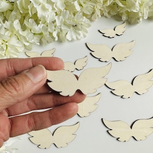 Op de afbeelding: Een verzameling kleine, onafgewerkte houten engelen vleugels. De vleugels zijn lichtbeige van kleur en hebben een glad oppervlak. Sommige vleugels zijn verspreid op een wit oppervlak, terwijl er één in een hand wordt gehouden. Witte bloemen op de achtergrond.