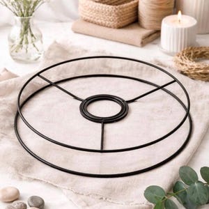 May include: A black metal lampshade frame with a circular design. The frame sits on a beige linen cloth, with decorative elements including a vase of white flowers, a candle, and twine in the background. The overall aesthetic is minimalist and natural.