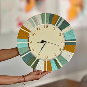 May include: A round clock with a white face and black hands. The clock is made of yarn in shades of green, blue, and yellow. The numbers on the clock are in a brown font.