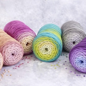 Può includere: Sei gomitoli di filato colorati nelle tonalità del rosa, viola, blu, verde e beige. Il filato è disposto in fila, con ogni gomitolo che mostra una sfumatura di colore. Piccole perline sono sparse intorno al filato.
