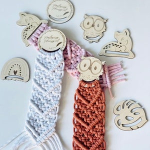 Op de afbeelding: Twee macramé wandhangers met houten uil- en kattentoppers. De wandhangers zijn wit en bruin met een roze en witte macramé hanger op de achtergrond. De houten toppers hebben de tekst "Reading Nook" erop.