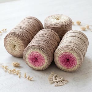 May include: Four cylindrical spools of yarn arranged in a cluster. Each spool features a gradient color transition, shifting from a deep pink center to a light cream, then to a soft brown, and finally to a darker brown shade on the outer edges.