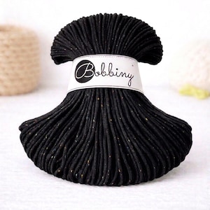Puede incluir: Ovillo de hilo negro con motas de color, envuelto con una etiqueta blanca con la palabra "Bobbing" en cursiva. El hilo está bien enrollado y parece suave, adecuado para proyectos de manualidades.