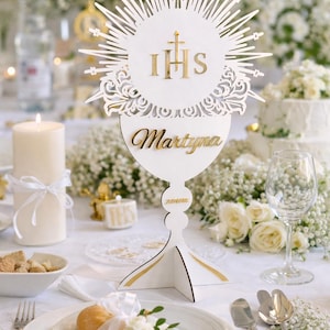 May include: A white laser-cut wooden table centerpiece with gold accents, featuring the letters IHS and the name "Martyna". The design includes a chalice shape and a radiant sunburst, suitable for a baptism or communion.
