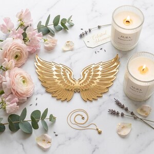 Op de afbeelding: Een goudkleurig metalen ornament in de vorm van een paar vleugels, vergezeld van een fijne gouden ketting, twee brandende kaarsen in geribbelde glazen potten met het label Aura, roze ranonkels, eucalyptus takjes en een label Celestial Adornments.