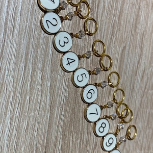 May include: Ten gold stitch markers with white round beads and clear crystal beads. Each marker has a white circle with a black number from 1 to 10.