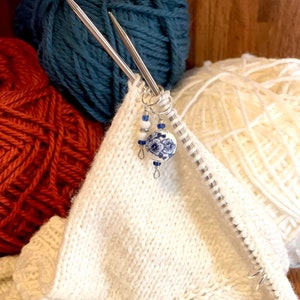 May include: White knitting needles with a blue and white ceramic bead charm. The needles are resting on a white knitted fabric. There are balls of yarn in the background, one is teal blue and the other is orange.